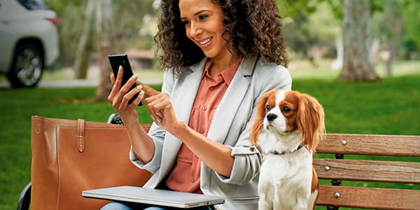 Chevrolet Divemotor - Mujer sentada en una plaza utilizando el servicio de onstar en su celular con su perro
